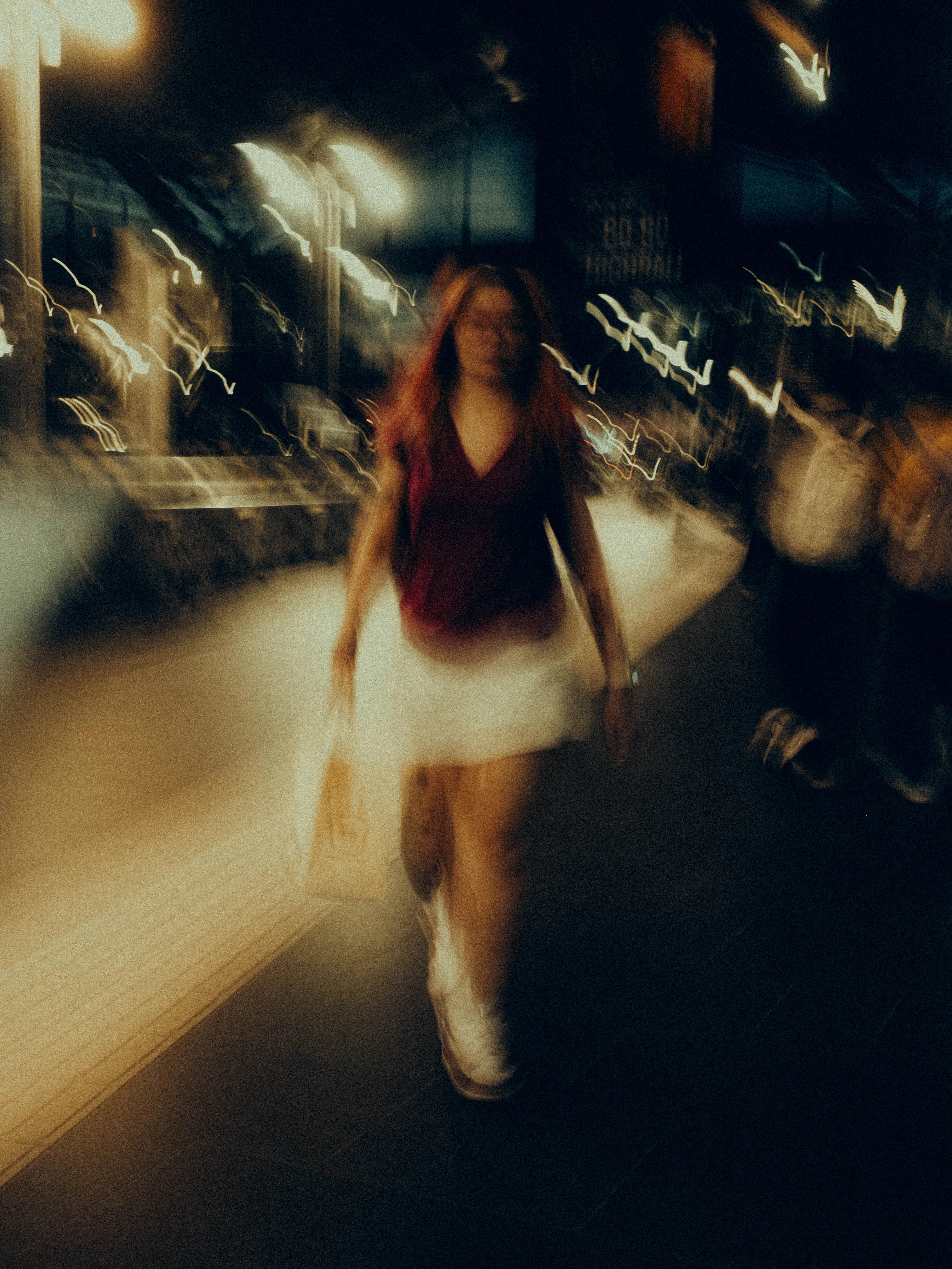 A red-haired woman in a dark top and white skirt walks alone through a warm night footpath, neon signs blooming into soft halos above the restless crowd dissolving behind her.