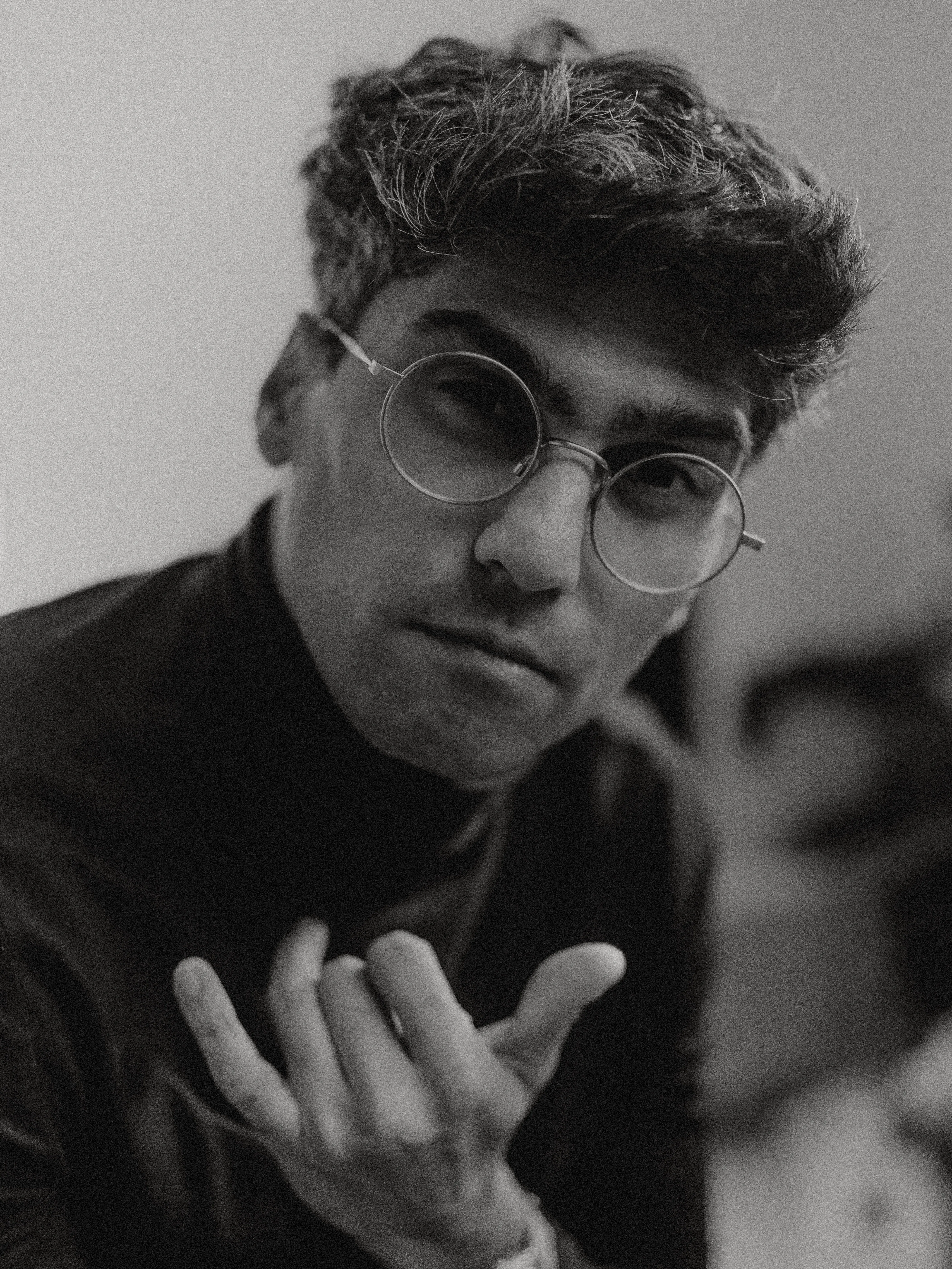 A black-and-white portrait of Abhay Desai in round wire-framed glasses and a dark high-necked top, chin resting against one hand, gazing directly at the camera in soft grainy light.